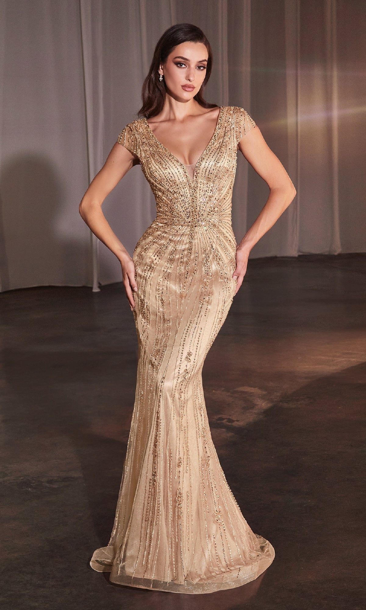 A woman stands in a dimly lit room wearing the Ladivine CZ0029, a long beaded formal dress featuring cap sleeves, a deep V-neckline, intricate beadwork, and a graceful mermaid skirt.